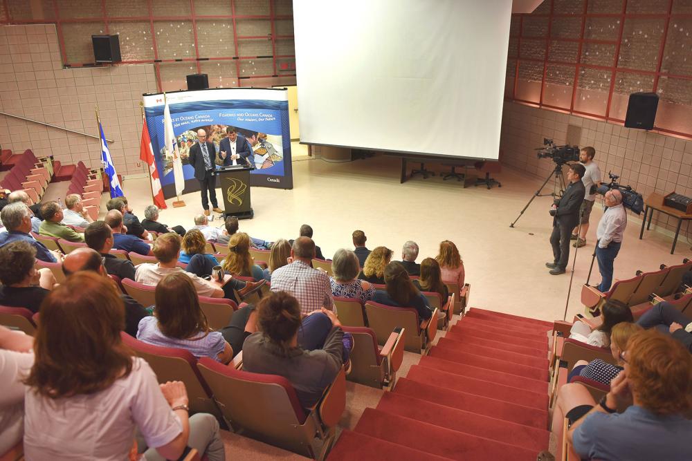 Photo d'un auditorium rempli de gens, avec le ministre LeBlanc et le député Rémi Massé au micro et des journalistes sur la scène