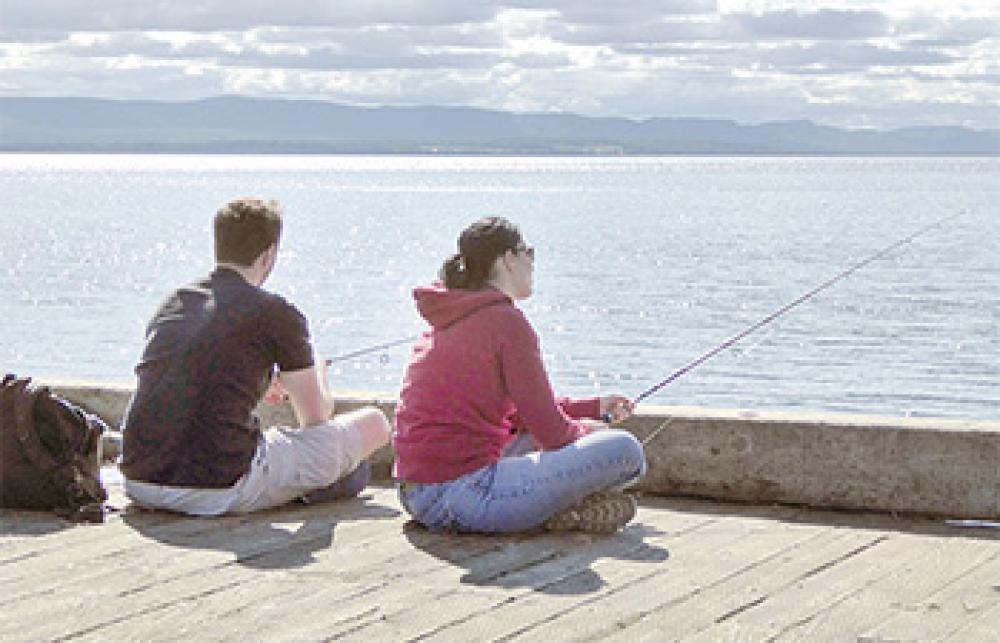 A man and a woman sitting on the edge of a dock are fishing by hand-line.